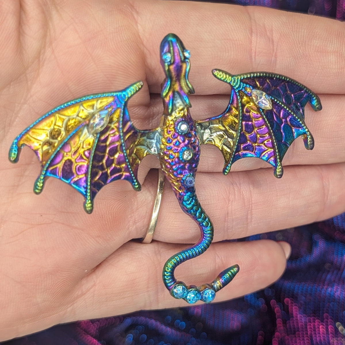 A small dragon-shaped brooch with iridescent enamel in blue, purple, green, and gold, displayed on a person's hand against a purple fabric background.