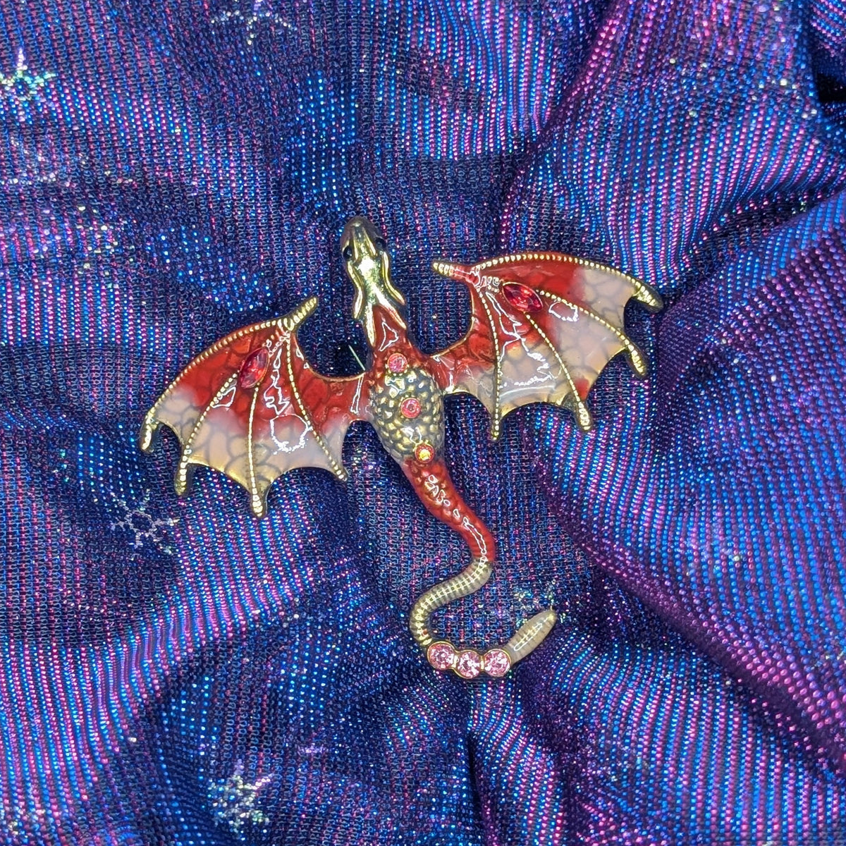 A dragon-shaped brooch with red wings and a jeweled body, placed on a shimmering purple and blue fabric background with snowflake motifs.