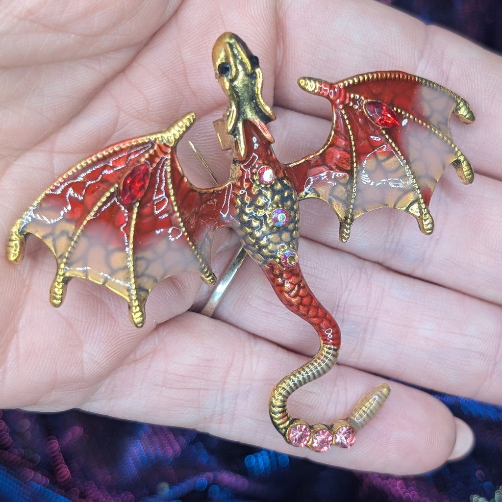 A small dragon figurine or brooch with gold-toned metal, red enamel, and translucent wings, resting on a person's hand against a purple and blue textured fabric background.
