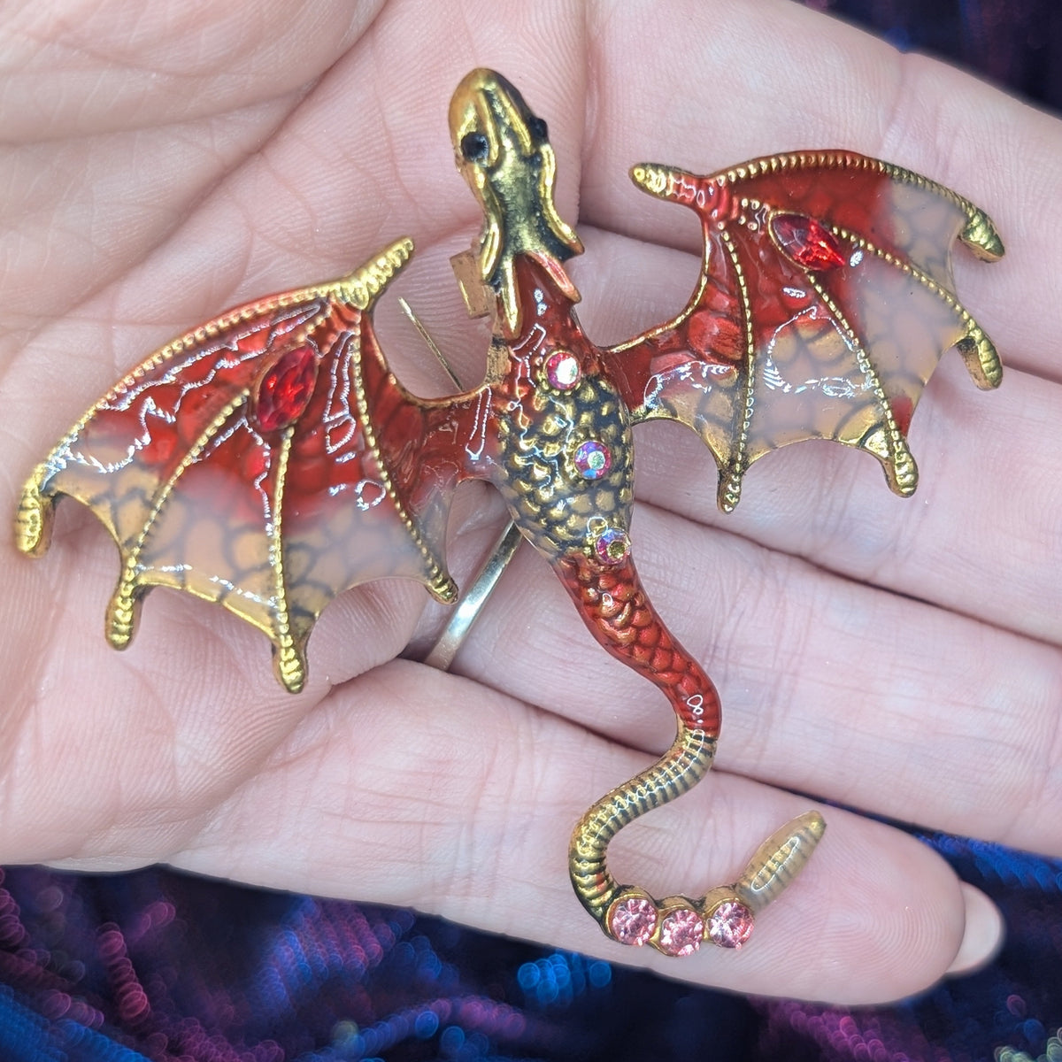 A small dragon figurine or brooch with gold-toned metal, red enamel, and translucent wings, resting on a person's hand against a purple and blue textured fabric background.