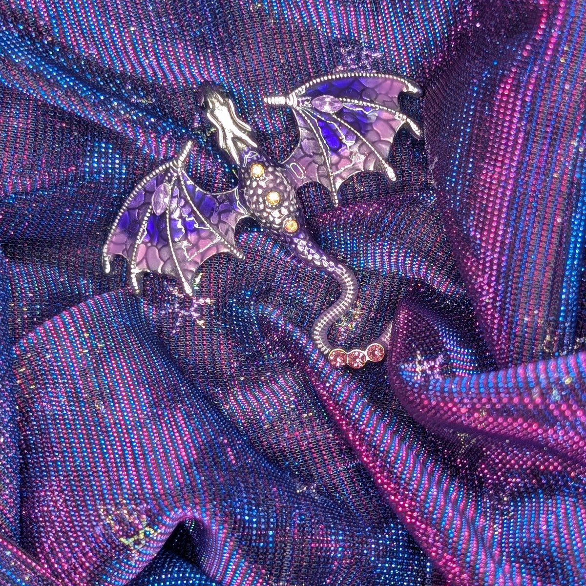 A detailed dragon brooch with purple wings and a silver body, resting on a shimmering purple and blue fabric background decorated with gold snowflakes.