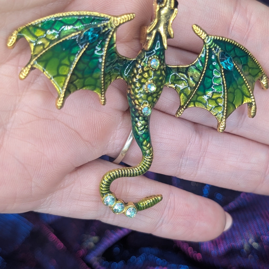 A close-up of a hand holding a detailed dragon brooch. The dragon is crafted with gold-toned metal and features vibrant green enamel scales, detailed wing membranes, and small clear rhinestones along its body and tail.