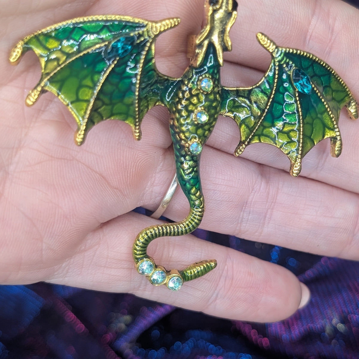 A close-up of a hand holding a detailed dragon brooch. The dragon is crafted with gold-toned metal and features vibrant green enamel scales, detailed wing membranes, and small clear rhinestones along its body and tail.