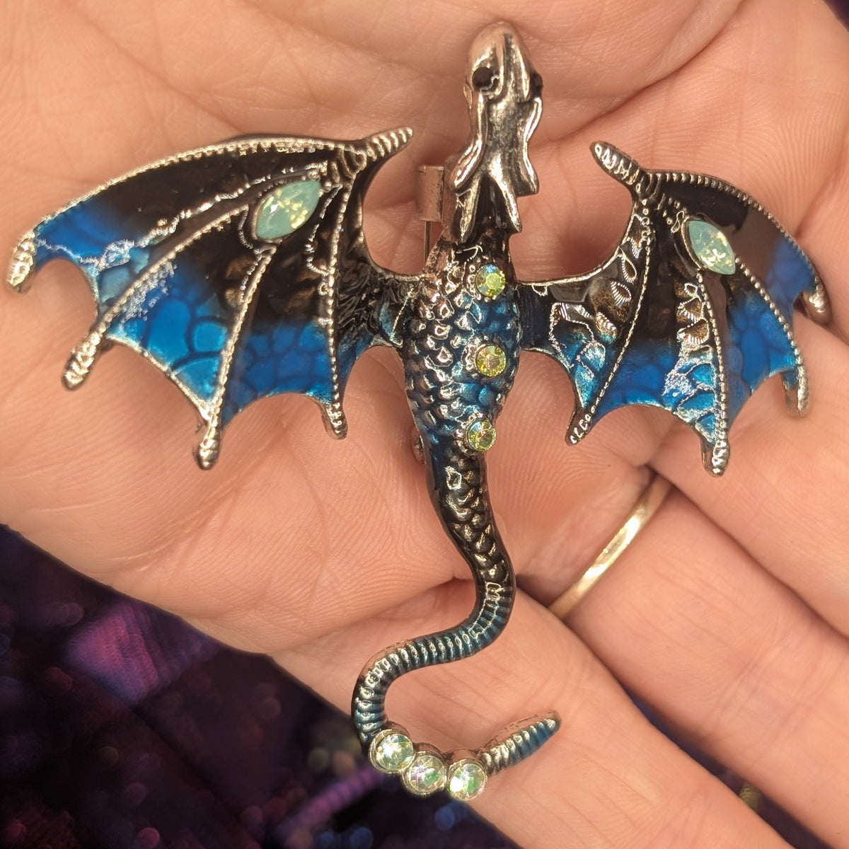 A detailed dragon pendant with blue enamel wings and body, silver-toned metal body and wings, and small green gemstones, held in a person's hand.